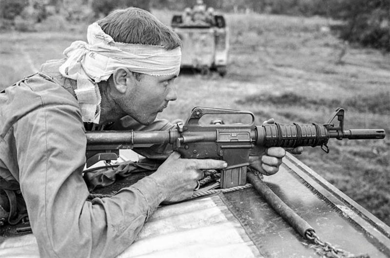 Pfc. Ron Fuller of Indianapolis, Ind., his head bandaged after being hit by shrapnel and his AR-15 at the ready, watches for the enemy from atop an armored personnel carrier.