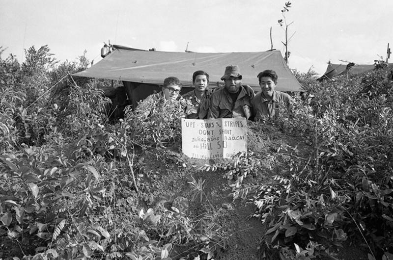 Stars and Stripes photographer John Olson, an unidentified man, UPI photographer William 'Bill' Hall, and UPI photographer Rikio Imajo (left to right) pose behind their tent on which they hung a sign reading "UPI, Stars and Stripes, Don't Shoot" in both English and Vietnamese.