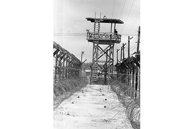 Army of the Republic of Vietnam Military Police (ARVN QC) guard stands atop a guard tower at Bien Hoa POW camp, overlooking the multiple barbed wire fences that surround the camp.
