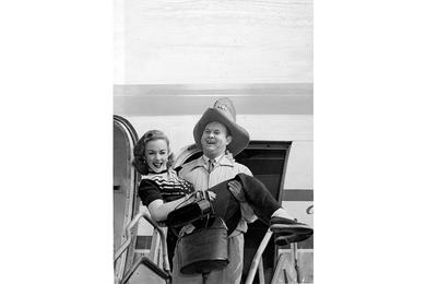 PRETTY BUNDLE -  Johnny Grant, USO troupe emcee, carries petite Piper Laurie from their Pan American Clipper at Tokyo 's International airport.
